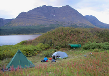 Load image into Gallery viewer, Camping scene with tents and equipment, electric bear fence, in a grassy field with mountains and a lake in the background.